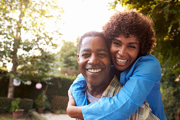 Woman and man smile at camera. The woman's arms are wrapped around him from behind.